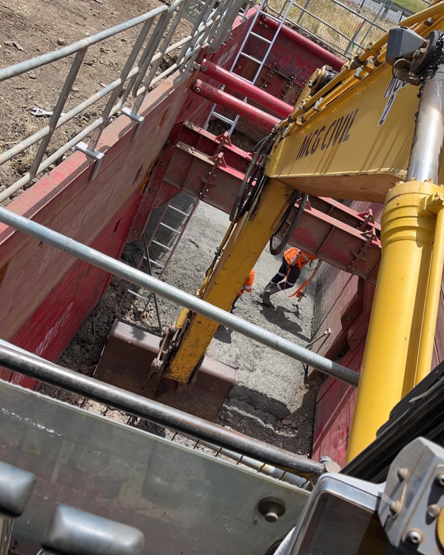 Excavator digging inside a red trench shield with a worker in hi-vis below