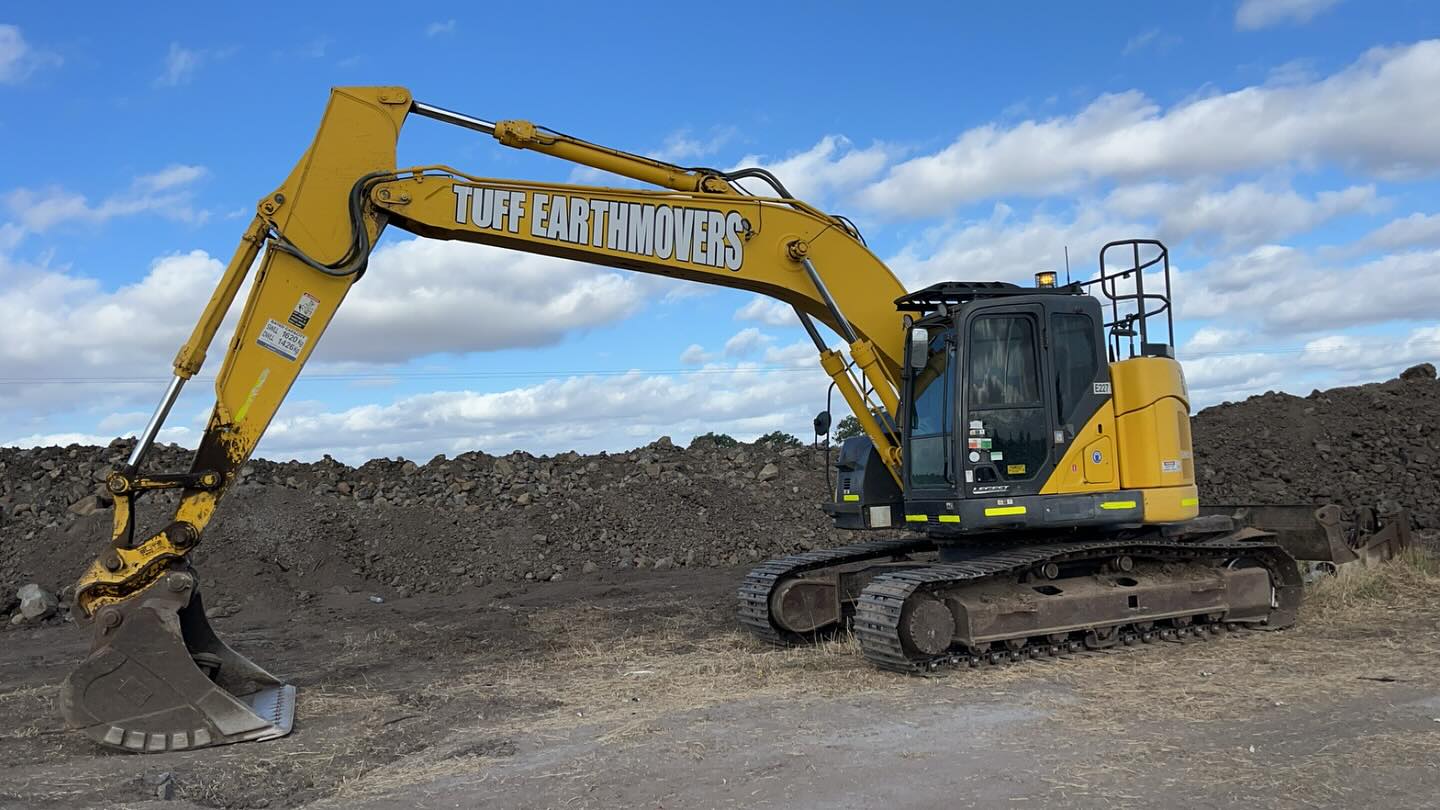 Yellow excavator lettered Tuff Earthmovers parked beside large dirt stockpiles