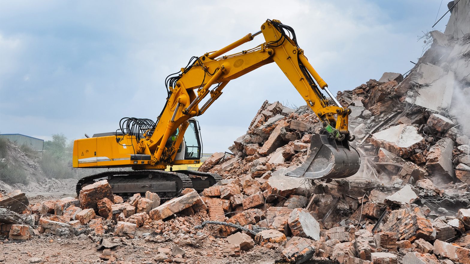Yellow excavator demolishing a brick building into a pile of rubble