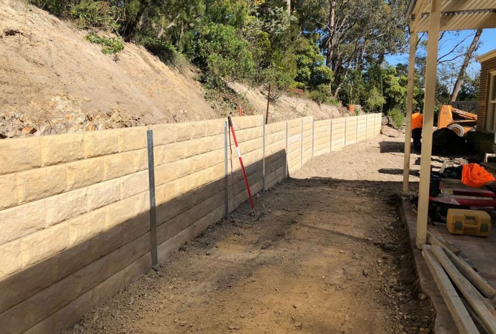 Sandstone-coloured block retaining wall with steel posts along a cut slope