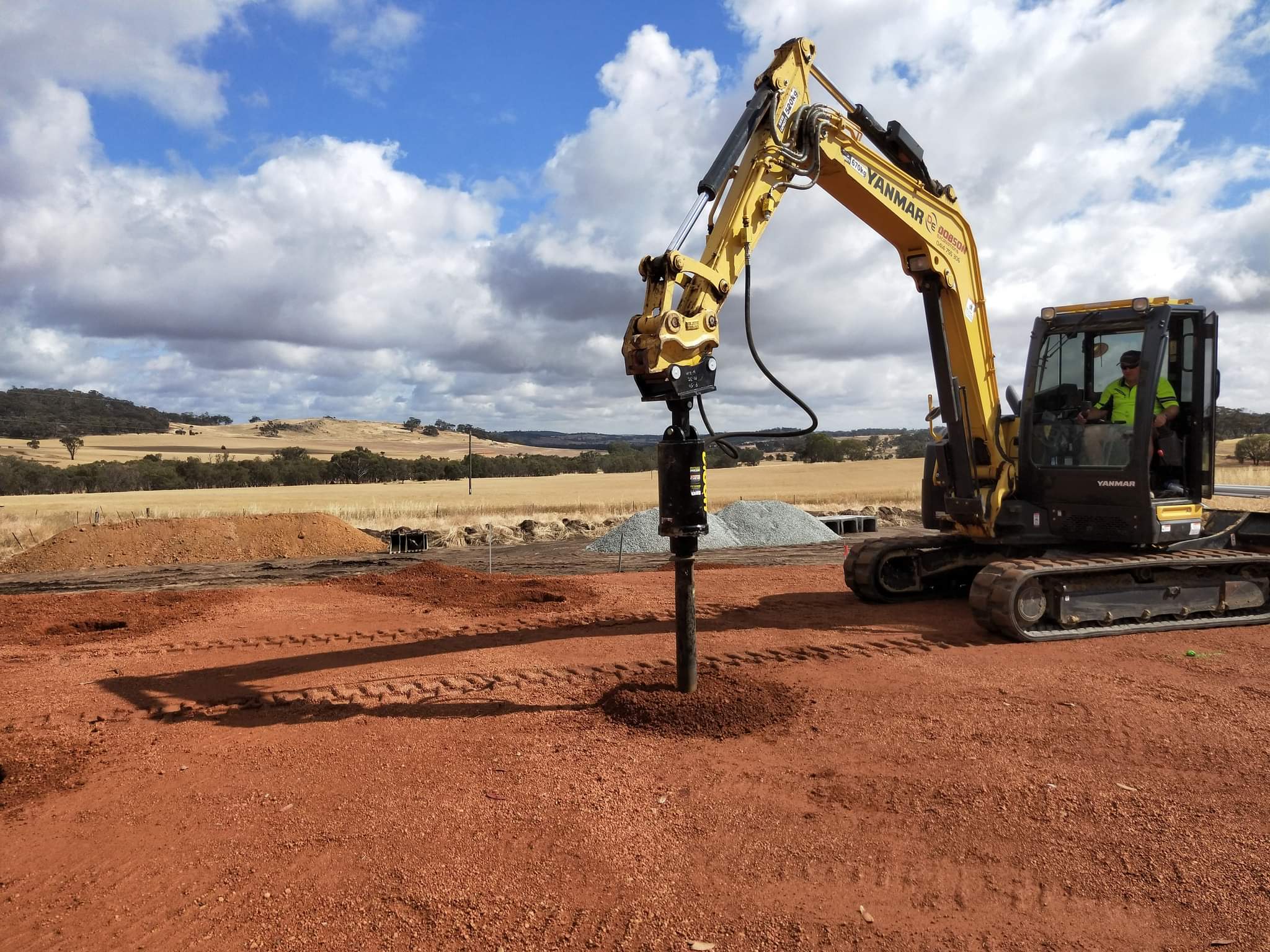Yellow excavator with an auger attachment drilling into red dirt